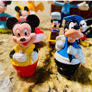 May include: A collection of colorful Mickey Mouse and Goofy figurines. Mickey Mouse is wearing a yellow shirt and red shorts. Goofy is wearing a blue graduation cap and gown. Other figures are visible in the background.