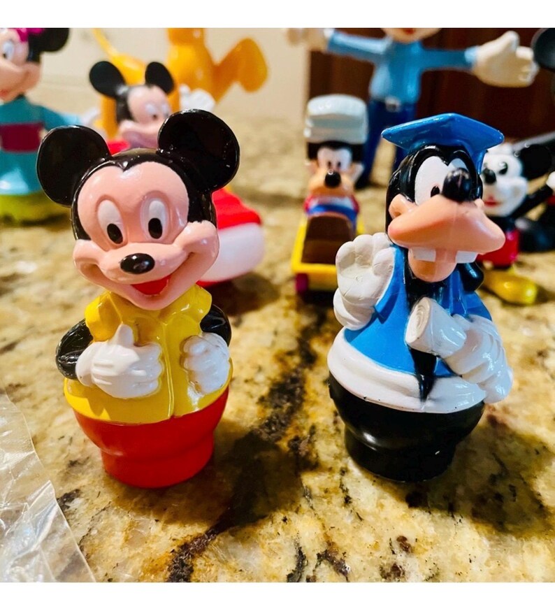 May include: A collection of colorful Mickey Mouse and Goofy figurines. Mickey Mouse is wearing a yellow shirt and red shorts. Goofy is wearing a blue graduation cap and gown. Other figures are visible in the background.