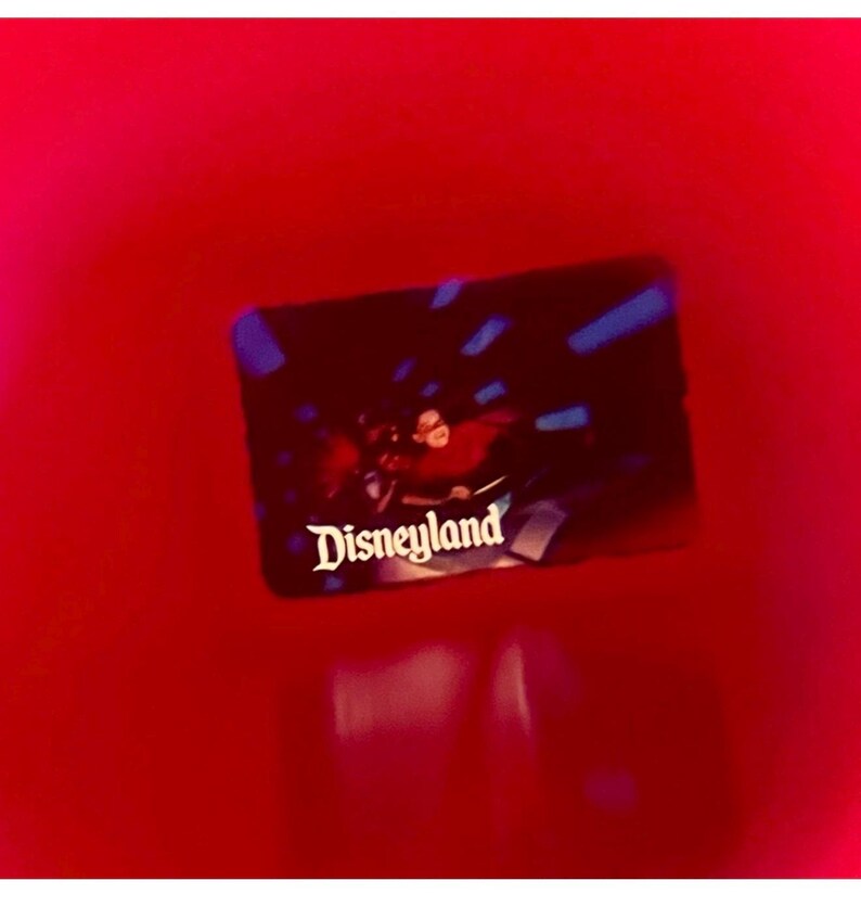 May include: A rectangular card with the word "Disneyland" in white script. The card has a dark background with a blurry image of people on a ride. The background is a bright red.