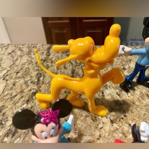 May include: A collection of Disney character figurines, including a bright yellow Pluto dog figure, a Mickey Mouse figure, and a Minnie Mouse figure with a pink bow. The figures are made of plastic and are set on a speckled countertop.