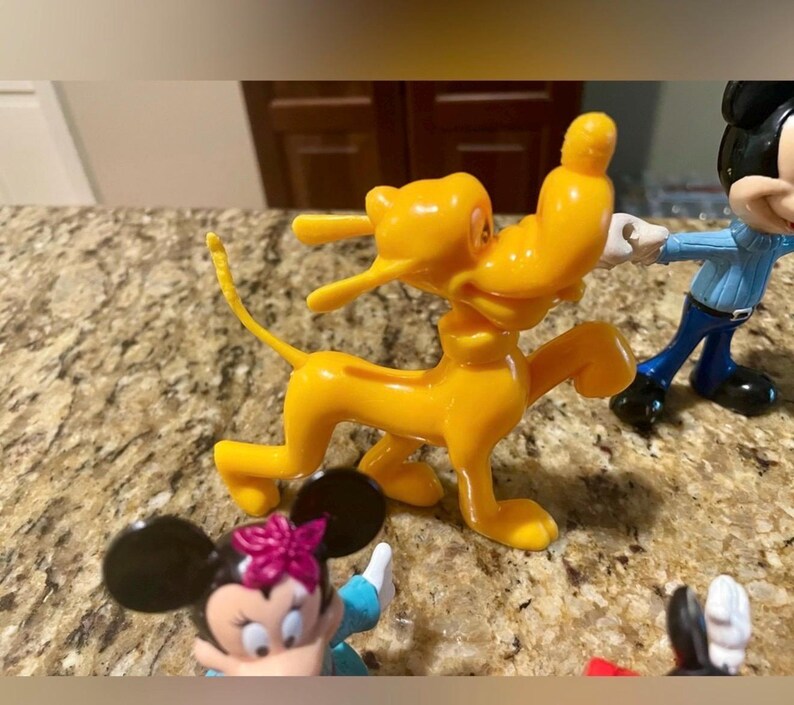 May include: A collection of Disney character figurines, including a bright yellow Pluto dog figure, a Mickey Mouse figure, and a Minnie Mouse figure with a pink bow. The figures are made of plastic and are set on a speckled countertop.