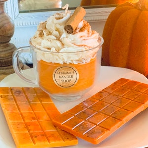 May include: A clear glass mug filled with orange wax, topped with white whipped wax and a cinnamon stick, featuring a round label that reads JASMINE'S KANDLE SHOP. Two rectangular orange wax bars with a grid pattern lie on the plate in front.