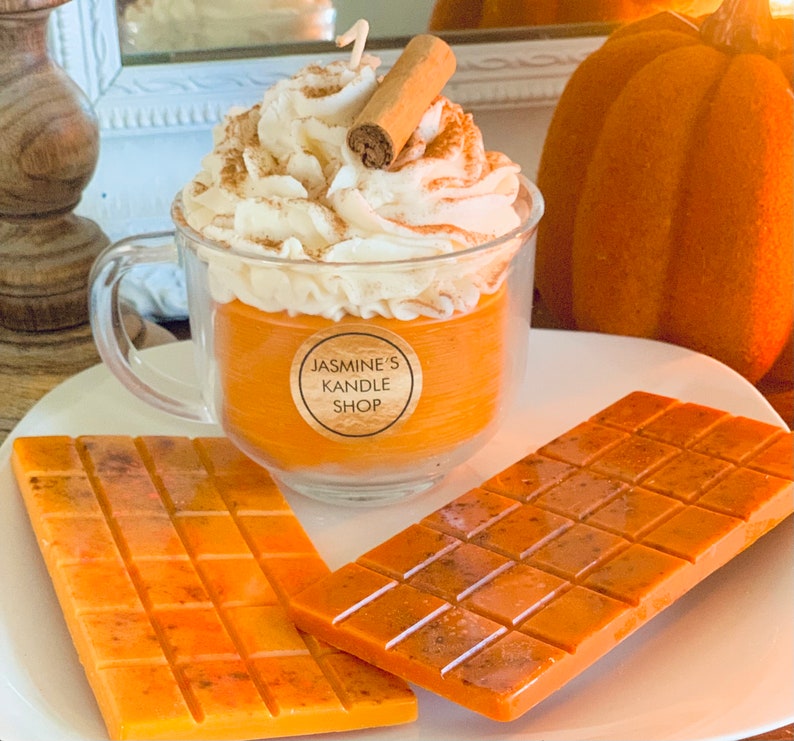 May include: A clear glass mug filled with orange wax, topped with white whipped wax and a cinnamon stick, featuring a round label that reads JASMINE'S KANDLE SHOP. Two rectangular orange wax bars with a grid pattern lie on the plate in front.