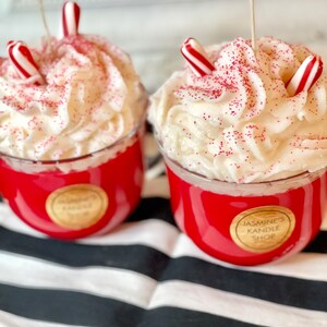 May include: Two red glass candles topped with white whipped wax resembling frosting, decorated with red sprinkles and small red and white striped candy cane pieces. Each candle features a round gold label reading Jasmine's Kandle Shop.