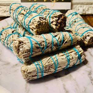 May include: A stack of white sage smudge sticks bundled with blue twine. The sticks are arranged in a pile on a white marble surface.