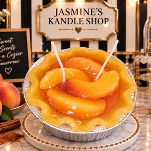 May include: A peach pie candle in a silver aluminum tin with a crimped edge. The candle features a glossy, golden-orange wax surface topped with realistic peach slices and two white wicks. The label reads JASMINE'S KANDLE SHOP.