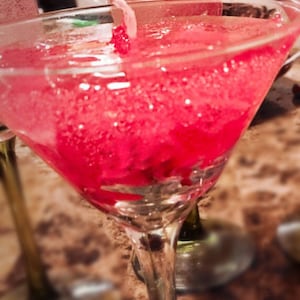 May include: A close-up of a pink, frosted, martini glass filled with a pink liquid. The glass is sitting on a table with other glasses out of focus in the background.