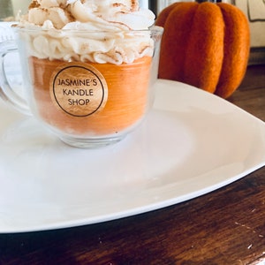 May include: A glass mug candle filled with orange wax, topped with white whipped wax and a cinnamon stick. A circular label on the front reads JASMINE'S KANDLE SHOP. The candle is placed next to a textured orange pumpkin decoration.
