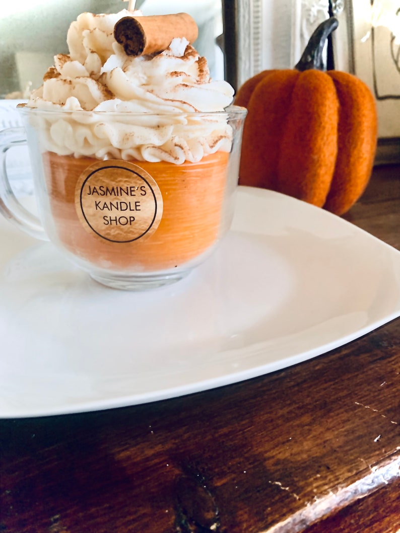 May include: A glass mug candle filled with orange wax, topped with white whipped wax and a cinnamon stick. A circular label on the front reads JASMINE'S KANDLE SHOP. The candle is placed next to a textured orange pumpkin decoration.