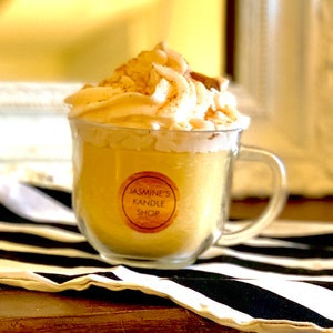 May include: A yellow candle in a clear glass mug topped with white wax shaped like whipped cream and a small cinnamon stick. A circular label on the front of the glass reads JASMINE'S KANDLE SHOP in black text on a tan background.