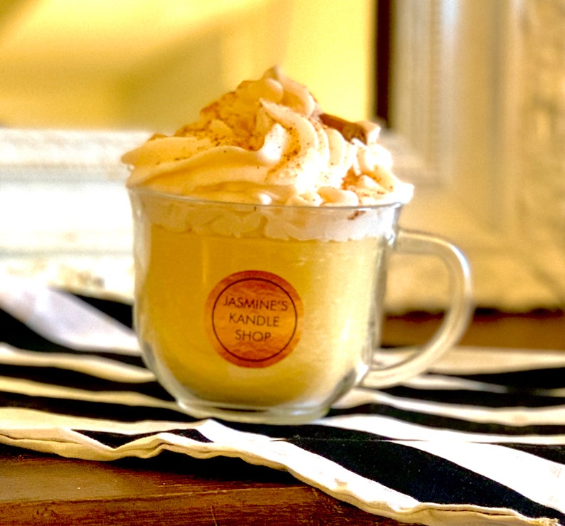 May include: A yellow candle in a clear glass mug topped with white wax shaped like whipped cream and a small cinnamon stick. A circular label on the front of the glass reads JASMINE'S KANDLE SHOP in black text on a tan background.