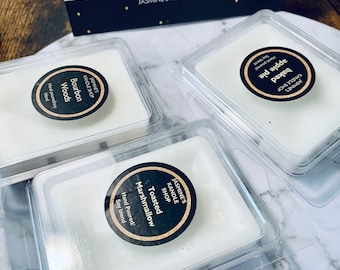 May include: Three small white wax melts in clear plastic containers. The wax melts are labeled with black and white circular stickers. The labels read "Toasted Marshmallow", "Bourbon Woods", and "Baked Apple Pie".