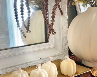 May include: Four white pumpkin-shaped candles on a gold tray. The candles are arranged in a row, with the largest candle in the center. The tray is on a wooden surface.