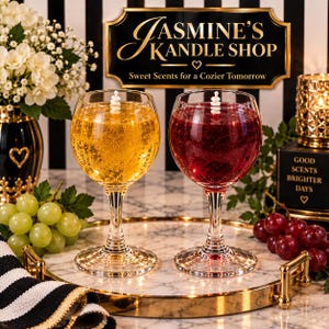 May include: Two wine glass candles, one filled with golden yellow wax and the other with deep red wax, each featuring a white wick. A sign reads JASMINE'S KANDLE SHOP Sweet Scents for a Cozier Tomorrow, with grapes and flowers nearby.