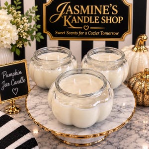 May include: Three white wax candles in clear, pumpkin-shaped glass jars with three wicks each. The candles are displayed on a round marble pedestal with a gold rim. A small black sign with white text reads Pumpkin Jar Candle with a heart symbol.