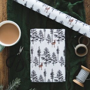 May include: A white wrapping paper with a black and brown deer and pine tree pattern. The paper is rolled up and lying on a green fabric surface. There is a cup of coffee, scissors, and twine on the surface.