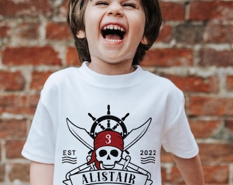 May include: A white short-sleeved t-shirt featuring a black and red graphic of a skull wearing a red bandana with the number 3, crossed swords, a ship's wheel, and the text EST 2022 ALISTAIR printed in a banner across the bottom centre.