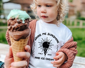 May include: A child wearing a white t-shirt with the text "Bring Me Sunshine and Ice Cream" in black. The shirt also features a black and white illustration of an ice cream cone with a sun behind it. The text "Italy 2023" is below the illustration. The child is holding a brown and green ice cream cone.