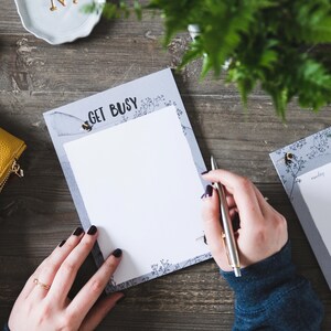 May include: A notepad with the words "GET BUSY" printed at the top, featuring a bee and floral design. A person is holding a silver pen over the blank white paper. The notepad is on a wooden surface.