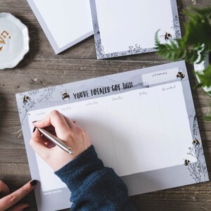 May include: A grey weekly planner notepad with the text "You've Totally Got This!" printed on the top. The notepad has a grey watercolour background with black and yellow bee illustrations. The days of the week are listed across the top: Monday, Tuesday, Wednesday, Thursday, Friday, and the weekend.
