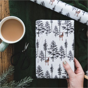 May include: White wrapping paper with a repeating pattern of black trees and brown deer. The paper is being held by a hand.