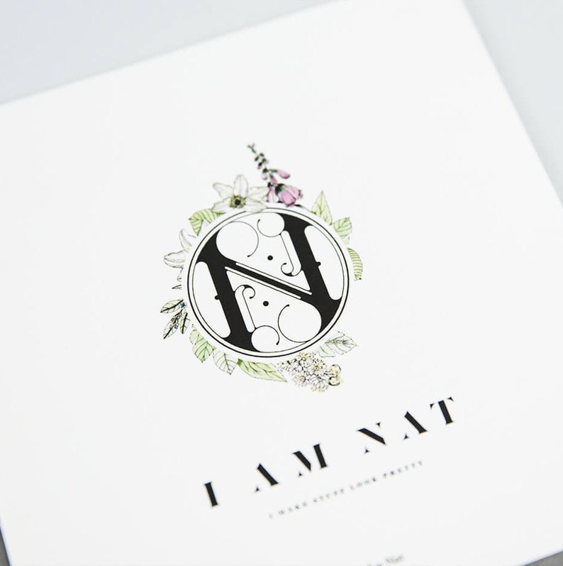 Pu&ograve; includere: Un monogramma in bianco e nero della lettera "H" circondato da una ghirlanda di foglie verdi e fiori rosa. Il testo "I AM NAT" &egrave; sotto il monogramma.