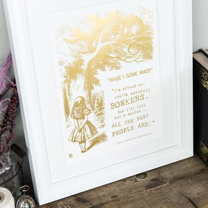 May include: Gold foil print of a scene from Alice in Wonderland with Alice standing in a forest and the Cheshire Cat in a tree above her. The quote reads "HAVE I GONE MAD?" "I'm afraid so. You're entirely BONKERS. But I'll tell you a secret... ALL THE BEST PEOPLE ARE."