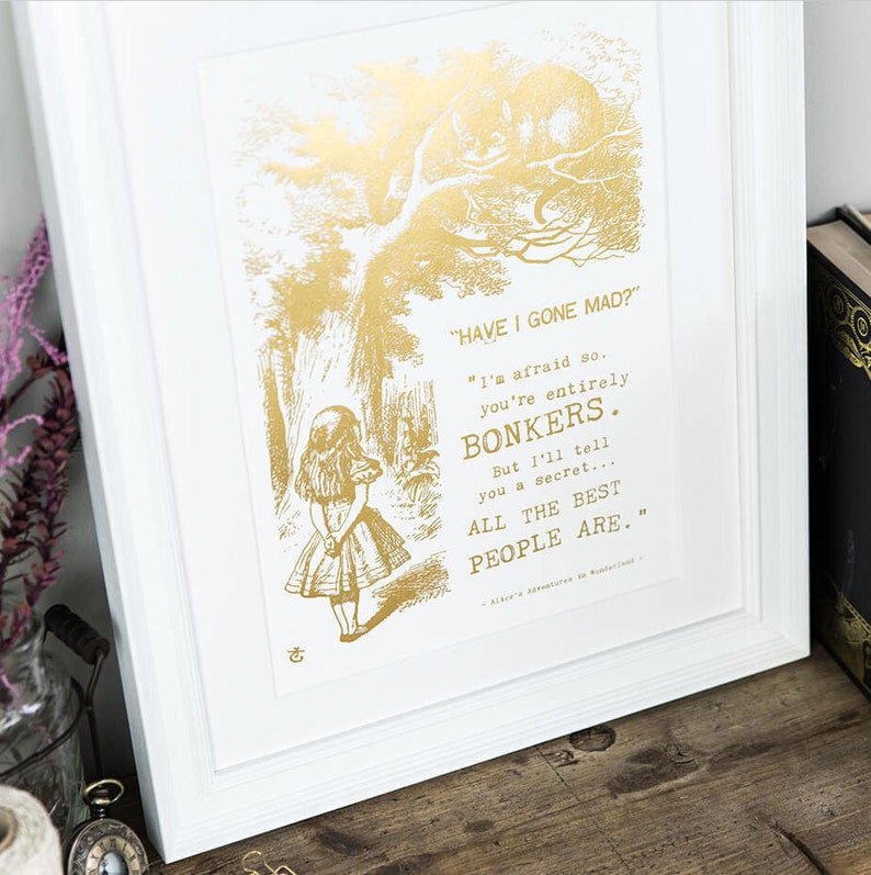 May include: Gold foil print of a scene from Alice in Wonderland with Alice standing in a forest and the Cheshire Cat in a tree above her. The quote reads "HAVE I GONE MAD?" "I'm afraid so. You're entirely BONKERS. But I'll tell you a secret... ALL THE BEST PEOPLE ARE."