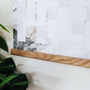 May include: A wooden calendar hanging on a white wall with a natural wood frame. The calendar has a white background with gray and brown abstract designs.