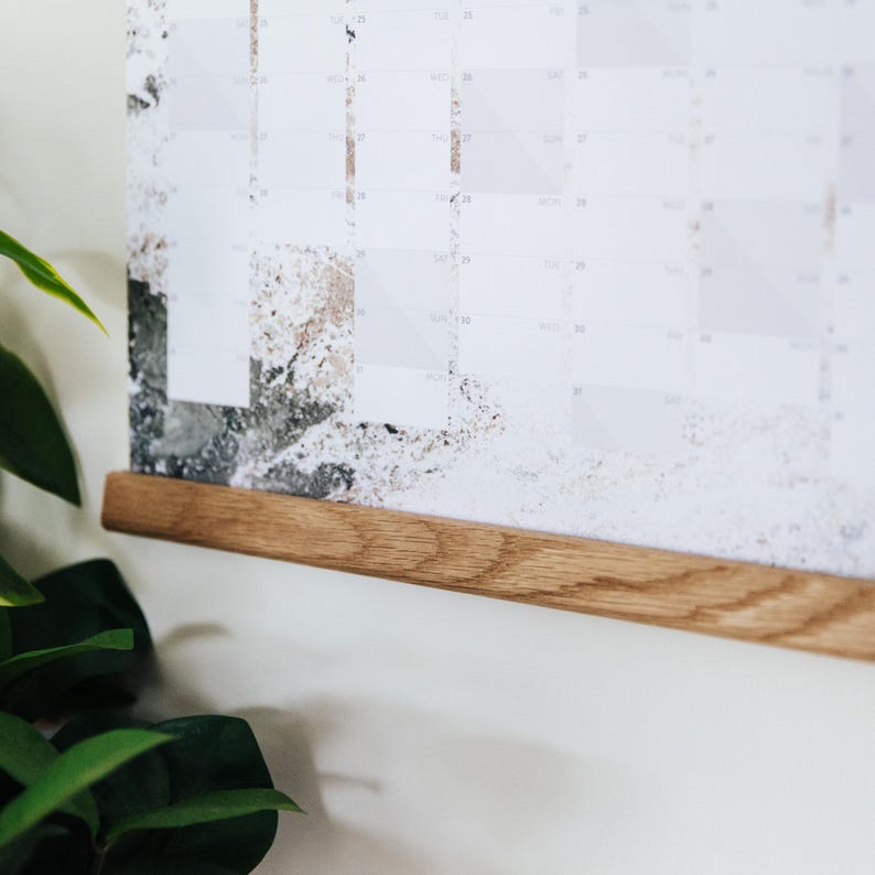 May include: A wooden calendar hanging on a white wall with a natural wood frame. The calendar has a white background with gray and brown abstract designs.