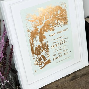 May include: A gold foil print of a quote from Alice in Wonderland, featuring a silhouette of Alice and the Cheshire Cat. The quote reads, "HAVE I GONE MAD?" "I'm afraid so. You're entirely BONKERS. But I'll tell you a secret... ALL THE BEST PEOPLE ARE..."