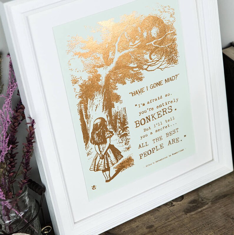 May include: A gold foil print of a quote from Alice in Wonderland, featuring a silhouette of Alice and the Cheshire Cat. The quote reads, "HAVE I GONE MAD?" "I'm afraid so. You're entirely BONKERS. But I'll tell you a secret... ALL THE BEST PEOPLE ARE..."