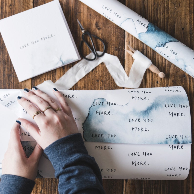 May include: A person wrapping a gift with blue and white wrapping paper that says "Love you more." The paper has a watercolour effect and the words are handwritten.