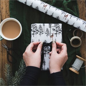 May include: A person tying a black ribbon on a white gift box with a forest and deer pattern. The gift box is wrapped in paper with a black and white design.