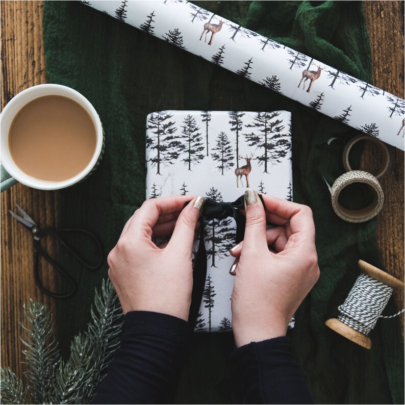 May include: A person tying a black ribbon on a white gift box with a forest and deer pattern. The gift box is wrapped in paper with a black and white design.