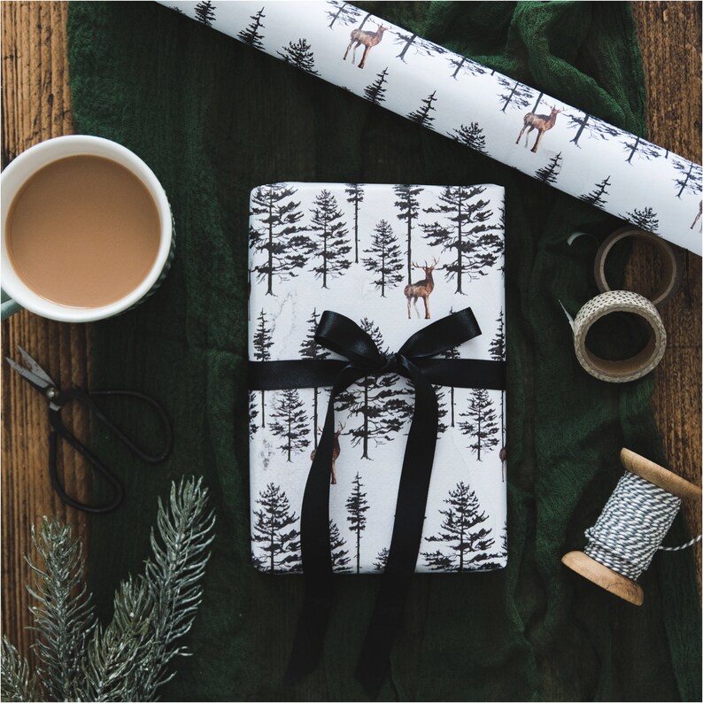 May include: A white gift box with a black ribbon and a forest and deer pattern. The box is wrapped in white paper with a black and white pattern of trees and deer. The box is tied with a black ribbon and is sitting on a green fabric surface.