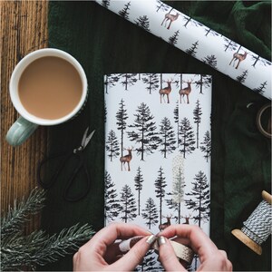 May include: A white wrapping paper with a repeating pattern of black trees and brown deer. The paper is being wrapped around a gift box.