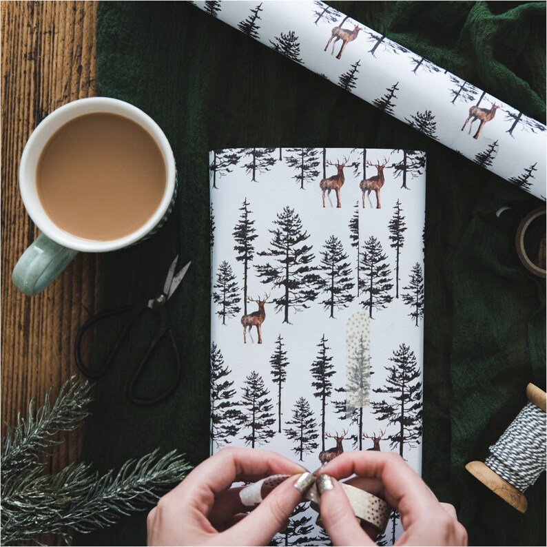 May include: A white wrapping paper with a repeating pattern of black trees and brown deer. The paper is being wrapped around a gift box.