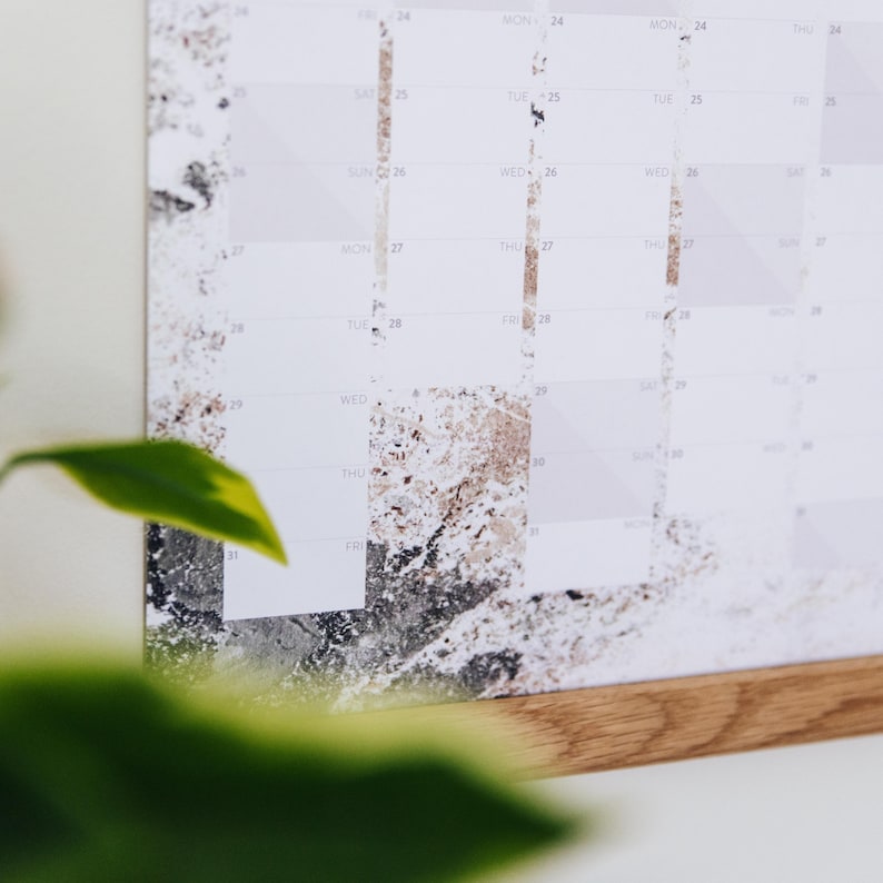 May include: A white calendar with a gray and brown marble pattern. The calendar is hanging on a wooden frame. The days of the week are labeled at the top of the calendar, and the numbers of the days are listed below. The calendar is for the month of August.