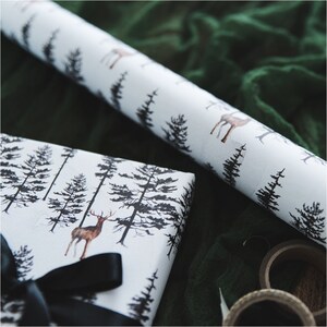 May include: White gift wrap with a black and brown deer and pine tree pattern. The gift wrap is rolled up and partially unrolled, revealing the pattern. A black ribbon is tied around the gift wrap.