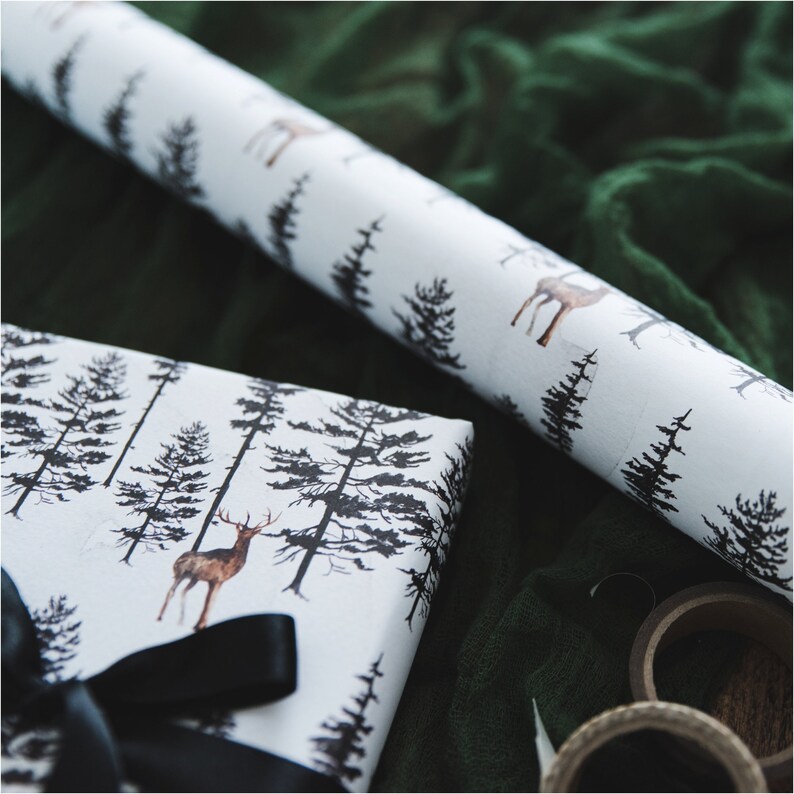May include: White gift wrap with a black and brown deer and pine tree pattern. The gift wrap is rolled up and partially unrolled, revealing the pattern. A black ribbon is tied around the gift wrap.