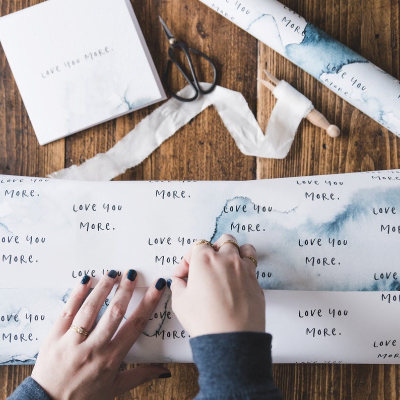 May include: A roll of white wrapping paper with a blue watercolour design and the words "Love you more" repeated throughout. A person is wrapping a gift with the paper.