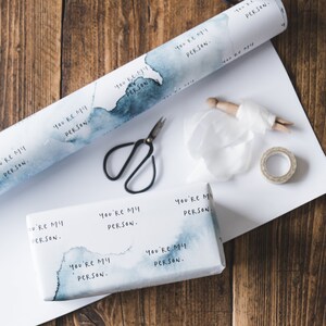 Puede incluir: Un rollo de papel de regalo azul y blanco con el texto "You're my person." impreso en &eacute;l. El papel est&aacute; enrollado y hay tijeras, cinta adhesiva y una pinza de madera sobre una superficie blanca.