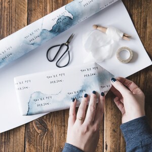 Puede incluir: Un papel de regalo blanco con dise&ntilde;os azules de acuarela y el texto "You're my person." repetido en todo el papel. Una persona est&aacute; envolviendo un regalo con el papel.