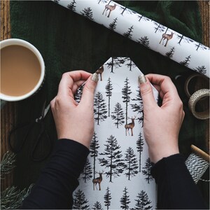 May include: A person wrapping a gift with white wrapping paper featuring a black and brown deer and pine tree pattern.