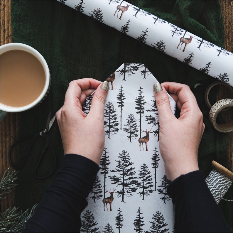 May include: A person wrapping a gift with white wrapping paper featuring a black and brown deer and pine tree pattern.