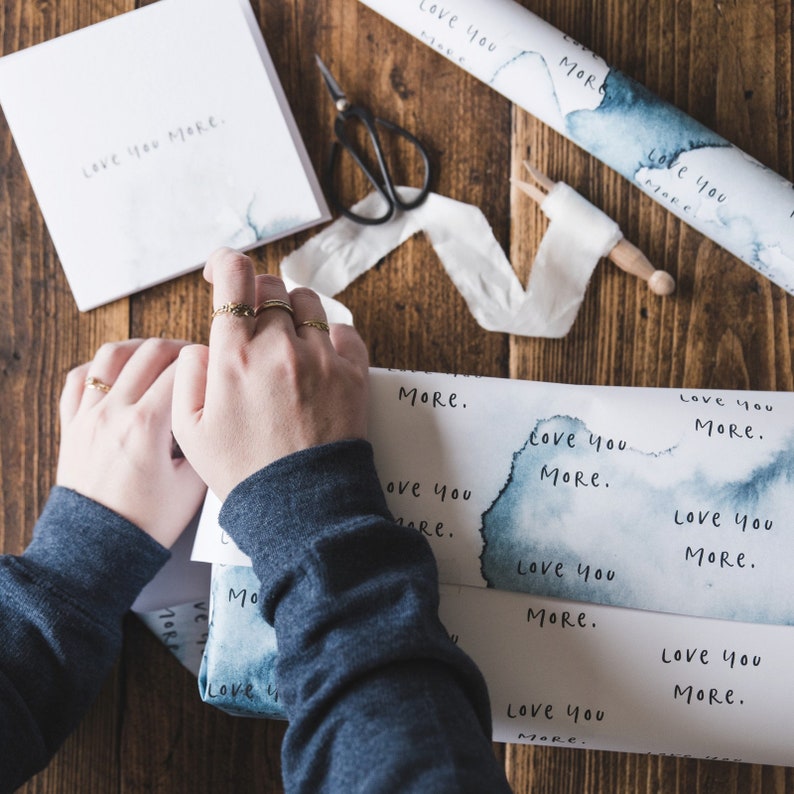 May include: A person wrapping a gift with blue and white watercolour wrapping paper that says "Love you more." The paper has a white background with blue watercolour splotches and the text is written in black.