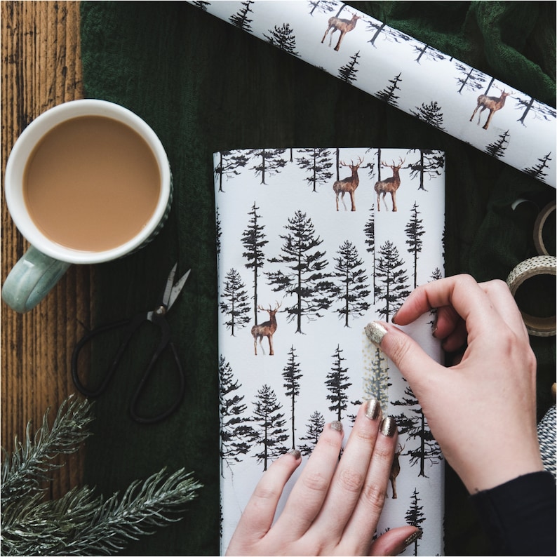 May include: A close-up of a person wrapping a gift with brown wrapping paper featuring a black and white pattern of deer and trees. The paper is being taped with a gold tape dispenser.