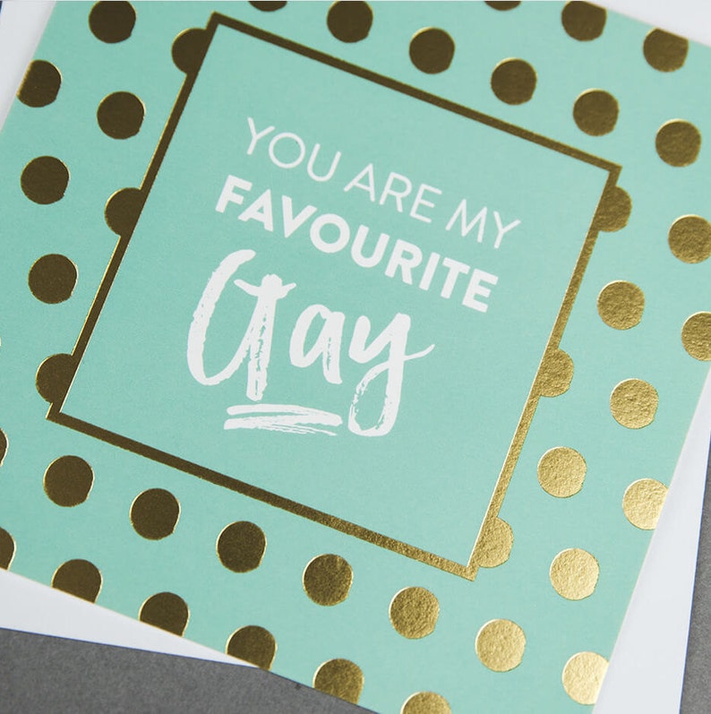 Pu&ograve; includere: Un biglietto di colore azzurro chiaro con pois dorati e un bordo dorato. Il biglietto dice "You are my favourite Gay" in testo bianco.
