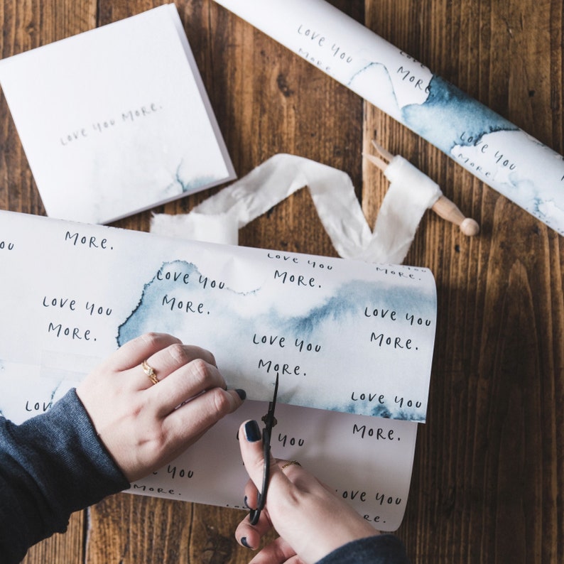 May include: A person is wrapping a gift with white wrapping paper featuring blue watercolour accents. The paper has the words "Love you more" printed on it. The person is using a pair of scissors to cut the paper.